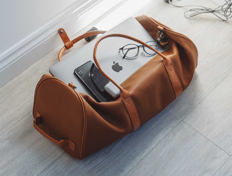 Reiserücktrittsversicherung nach Buchung: Gepackte Ledertasche, MacBook, iPhone und Kopfhörer liegen auf dem Holzboden